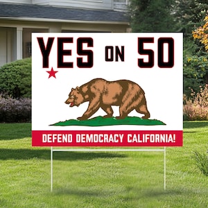 May include: A white yard sign with the text "YES ON 50" in black and red, featuring a brown bear graphic and the words "DEFEND DEMOCRACY CALIFORNIA!" in red. The sign is displayed on a green lawn in front of a house.