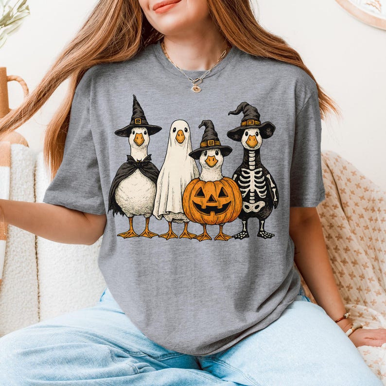 May include: Gray t-shirt featuring a Halloween-themed graphic. The design showcases four geese dressed in costumes: a witch, a ghost, a jack-o'-lantern, and a skeleton. The geese are wearing black hats and capes. The graphic is in a cartoon style.
