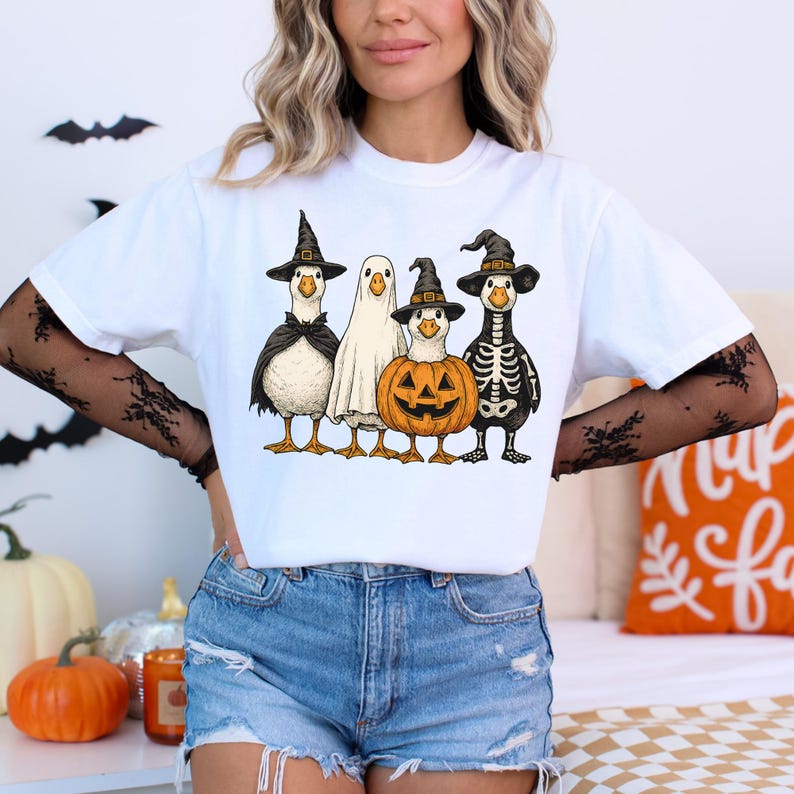 May include: White t-shirt featuring a Halloween-themed graphic. The design includes five cartoon geese dressed in costumes: a witch, a ghost, a pumpkin, and a skeleton. The geese are standing in a row, with a black bat silhouette in the background.