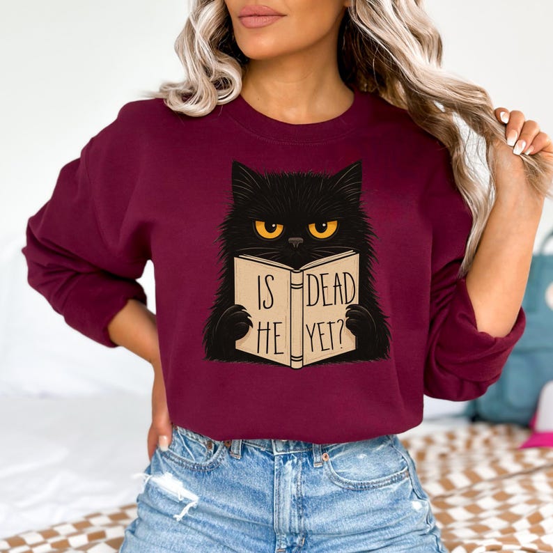 Pode incluir: Camisola bord&ocirc; com um gato preto lendo um livro. O livro diz "IS HE DEAD YET?". O gato tem olhos amarelos. A camisola &eacute; de cor s&oacute;lida e de estilo casual.