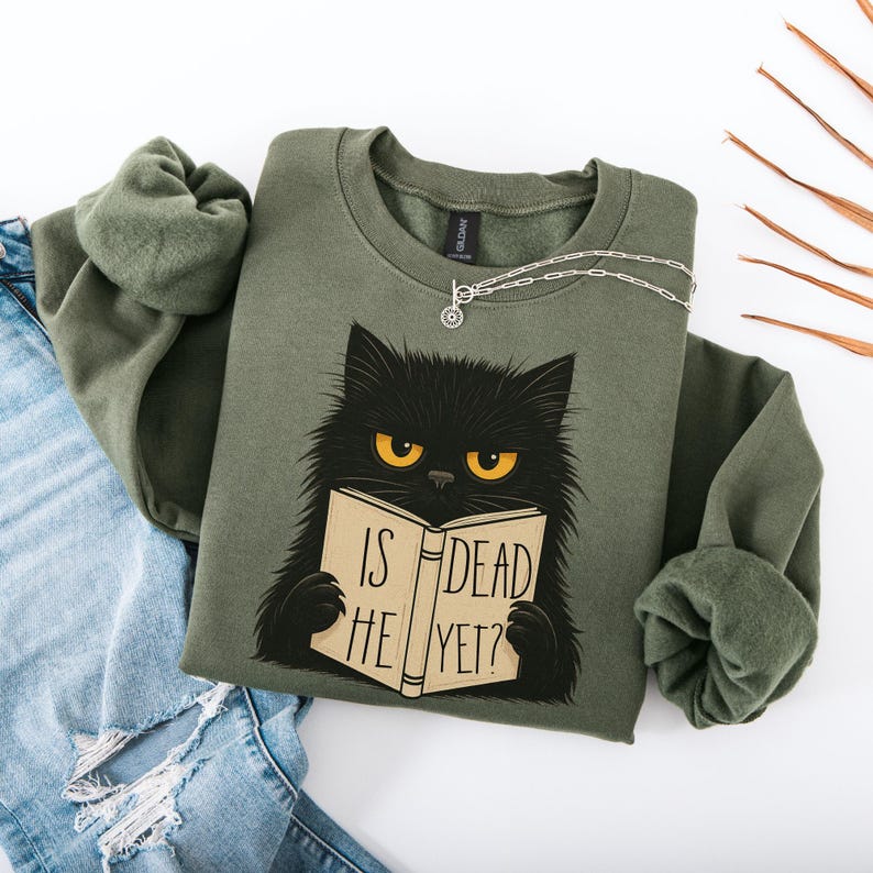Pode incluir: Um moletom verde-oliva com um gato preto lendo um livro. O livro diz "IS HE DEAD YET?". O gato tem olhos amarelos. Um colar de prata est&aacute; sobre o moletom, que &eacute; combinado com jeans azuis.