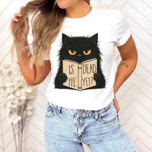 May include: White t-shirt featuring a black cat with yellow eyes reading a book. The book's cover reads "IS HE DEAD YET?" in black text. The cat has a fluffy appearance and is centered on the shirt.
