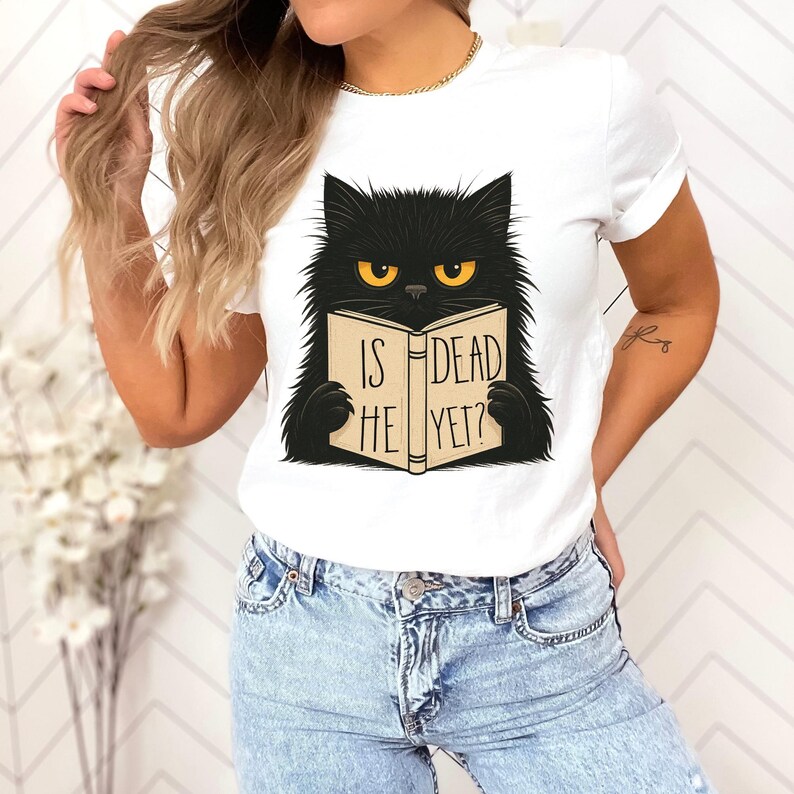 May include: White t-shirt featuring a black cat with yellow eyes reading a book. The book's cover reads "IS HE DEAD YET?" in black text. The cat has a fluffy appearance and is centered on the shirt.
