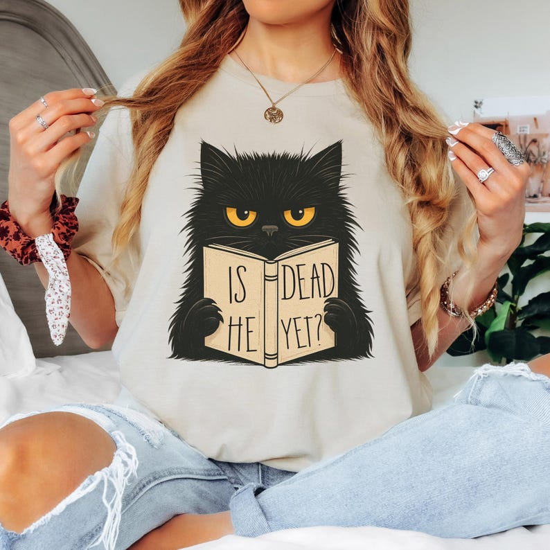 May include: Beige t-shirt featuring a black cat reading a book with the text "IS HE DEAD YET?" The cat has yellow eyes and a stern expression. The shirt is a casual style.