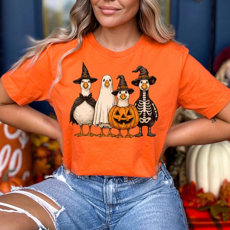 May include: Orange t-shirt featuring a Halloween-themed graphic. The graphic depicts four cartoon ducks dressed in costumes: a witch, a ghost, a jack-o'-lantern, and a skeleton. The ducks are standing in a row. The shirt is made of a soft material.