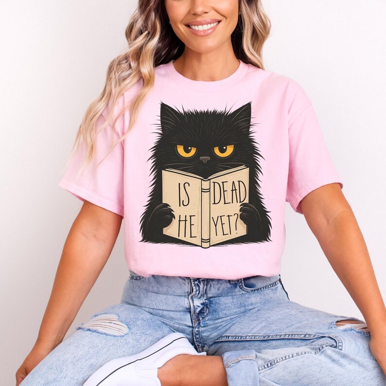 May include: A light pink t-shirt featuring a black cat illustration with yellow eyes. The cat is holding a book with the text "IS HE DEAD YET?" printed on the cover. The shirt is a casual wear item.