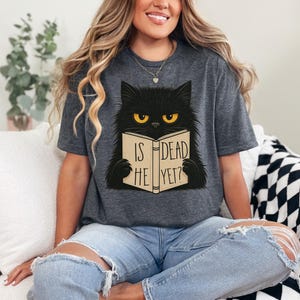 May include: Dark gray t-shirt featuring a black cat with yellow eyes reading a book. The book's cover reads "IS HE DEAD YET?" The cat is the central graphic on the shirt.