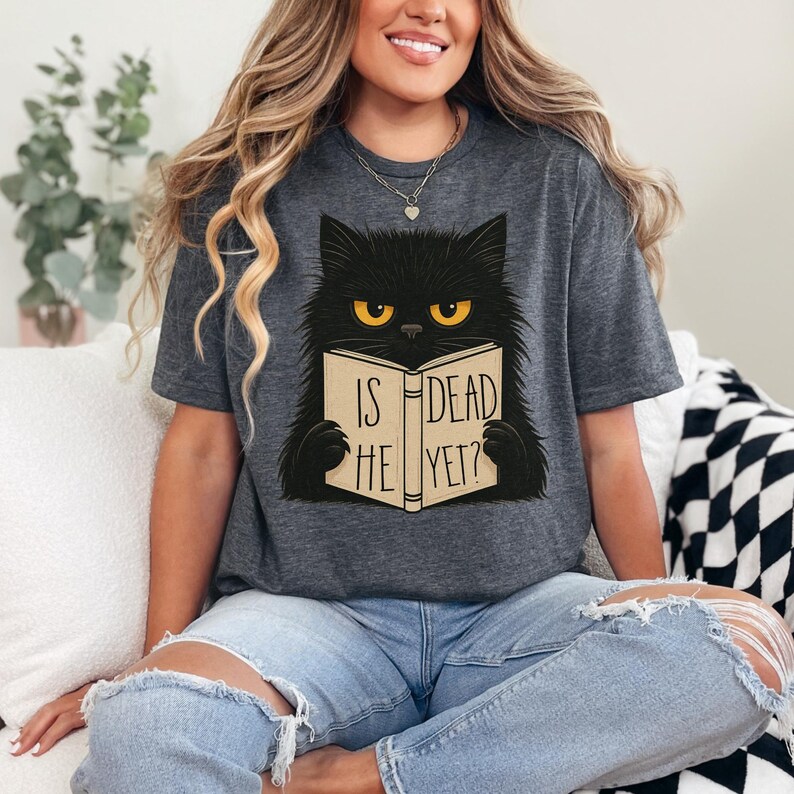 May include: Dark gray t-shirt featuring a black cat with yellow eyes reading a book. The book's cover reads "IS HE DEAD YET?" The cat is the central graphic on the shirt.