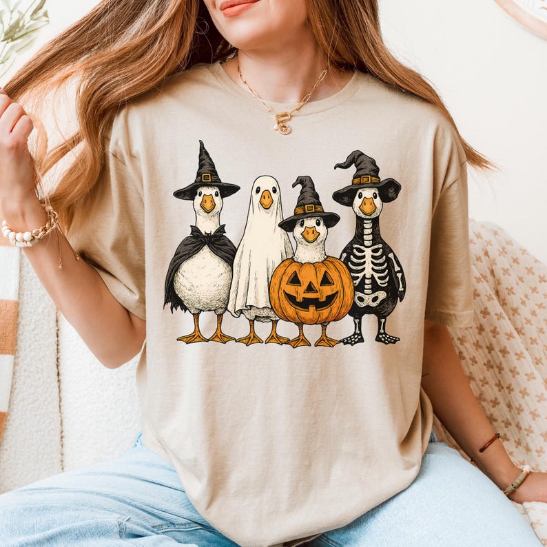 May include: Beige t-shirt featuring a Halloween-themed graphic. The design includes four cartoon ducks dressed in costumes: a witch, a ghost, a jack-o'-lantern, and a skeleton. The ducks are standing in a row, with the witch duck on the left.