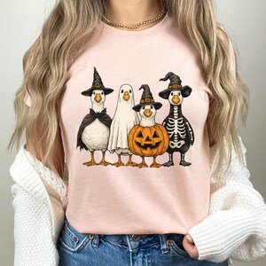 May include: A light pink t-shirt featuring a Halloween-themed graphic. The graphic depicts four geese dressed in Halloween costumes: a witch, a ghost, a jack-o'-lantern, and a skeleton. The geese are wearing black hats and capes.