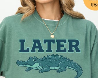 May include: Light green t-shirt with the word "LATER" in dark blue block letters above a cartoon alligator. The alligator is light blue with darker blue details. The shirt is a relaxed fit.