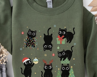 May include: Military green sweatshirt with a Christmas-themed design of black cats. The cats are illustrated in different poses, some with festive accessories like Santa hats and reindeer antlers. The design includes Christmas lights and gifts, creating a playful seasonal look.
