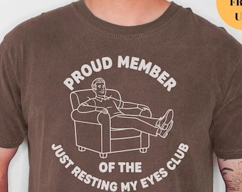 May include: Espresso-colored t-shirt with a white graphic. The graphic reads "PROUD MEMBER OF THE JUST RESTING MY EYES CLUB" and features a person reclining in a chair. The shirt is a crew neck.