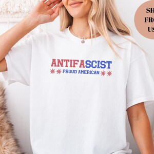 May include: White t-shirt with the text "ANTIFASCIST" in red and blue, with "PROUD AMERICAN" below. The design includes star graphics. The shirt is a casual style.