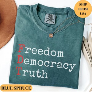 Puede incluir: Camiseta azul abeto con las palabras "Freedom, Democracy, Truth" impresas en blanco y rojo. Un collar de oro con un colgante descansa sobre la camisa. La camisa est&aacute; doblada y tiene una etiqueta Comfort Colors.