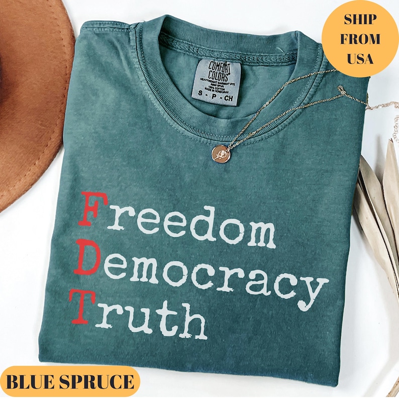 Puede incluir: Camiseta azul abeto con las palabras "Freedom, Democracy, Truth" impresas en blanco y rojo. Un collar de oro con un colgante descansa sobre la camisa. La camisa est&aacute; doblada y tiene una etiqueta Comfort Colors.