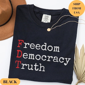 Puede incluir: Camiseta negra con las palabras "Freedom Democracy Truth" impresas en blanco y rojo. Tambi&eacute;n se ven un collar dorado y un sombrero marr&oacute;n. La camiseta es de material Comfort Colors.