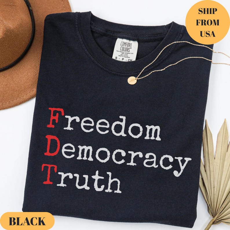 Puede incluir: Camiseta negra con las palabras "Freedom Democracy Truth" impresas en blanco y rojo. Tambi&eacute;n se ven un collar dorado y un sombrero marr&oacute;n. La camiseta es de material Comfort Colors.