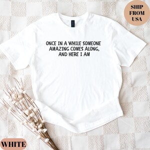 May include: White t-shirt with the text "ONCE IN A WHILE SOMEONE AMAZING COMES ALONG, AND HERE I AM" in black. The shirt is a crew neck style. The words "WHITE" and "SHIP FROM USA" are also visible.