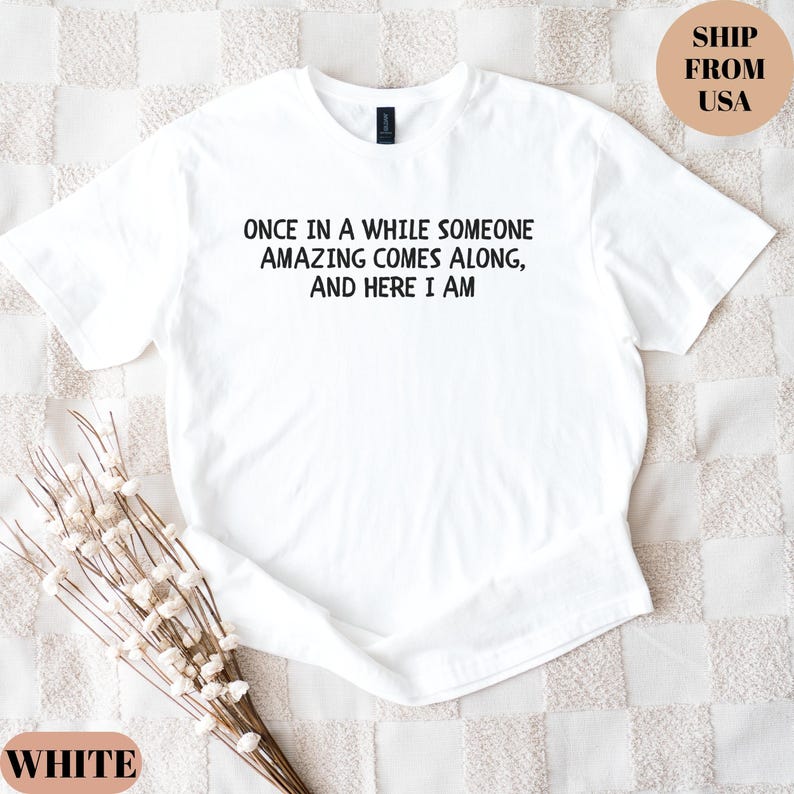 May include: White t-shirt with the text "ONCE IN A WHILE SOMEONE AMAZING COMES ALONG, AND HERE I AM" in black. The shirt is a crew neck style. The words "WHITE" and "SHIP FROM USA" are also visible.