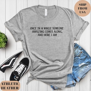 May include: Athletic heather gray t-shirt with the text "ONCE IN A WHILE SOMEONE AMAZING COMES ALONG, AND HERE I AM." The shirt is paired with denim shorts and black and white sneakers. The shirt is made of a soft material.
