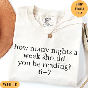Puede incluir: Camiseta blanca con el texto "how many nights a week should you be reading? 6-7". La camiseta tiene cuello redondo y mangas cortas. Un collar dorado con un colgante est&aacute; drapeado sobre la camiseta. Un sombrero marr&oacute;n es visible en el fondo.