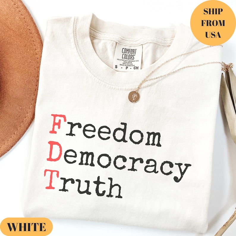 Puede incluir: Camiseta color crema con las palabras "Freedom Democracy Truth" impresas en negro y rojo. Un collar dorado con un colgante descansa sobre la camiseta. La camiseta tiene una etiqueta que dice "Comfort Colors".