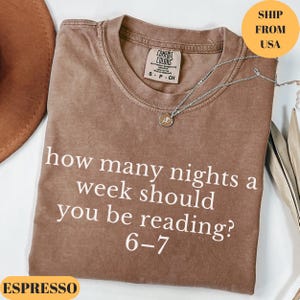 Puede incluir: Camiseta color espresso con el texto blanco "how many nights a week should you be reading? 6-7". La camiseta tiene cuello redondo y una peque&ntilde;a etiqueta. Un collar plateado con un colgante est&aacute; sobre la camiseta.