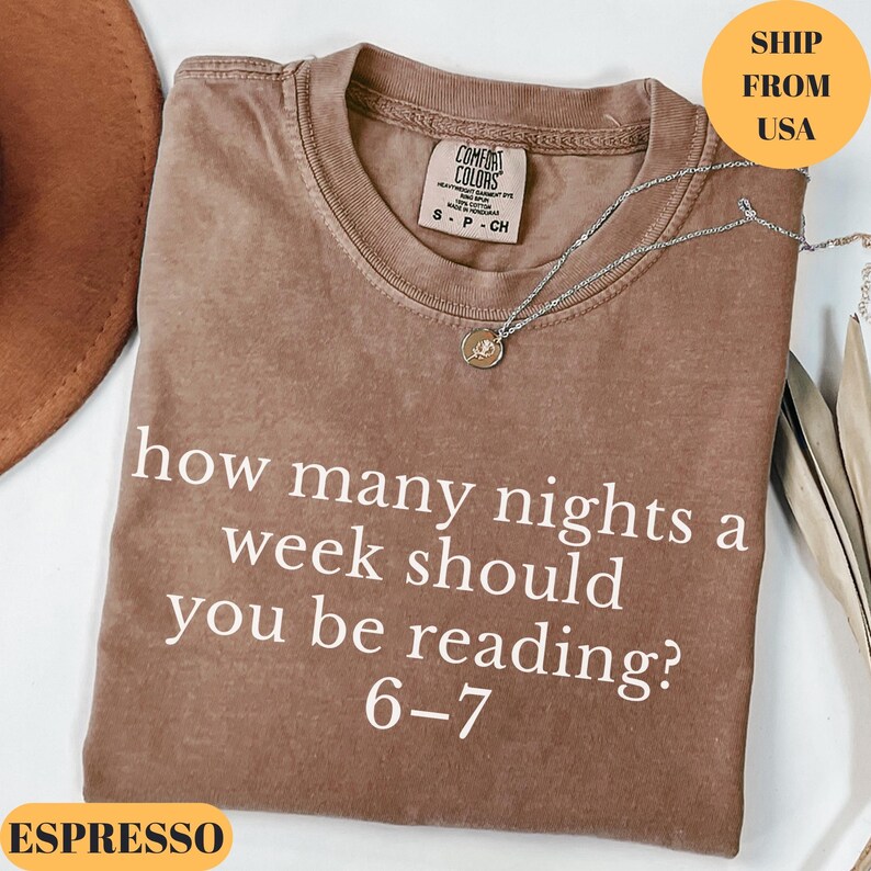 Puede incluir: Camiseta color espresso con el texto blanco "how many nights a week should you be reading? 6-7". La camiseta tiene cuello redondo y una peque&ntilde;a etiqueta. Un collar plateado con un colgante est&aacute; sobre la camiseta.
