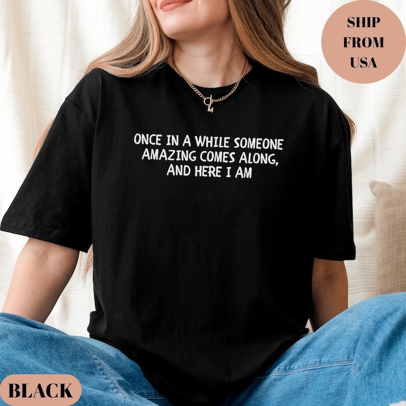 May include: Black t-shirt with the text "ONCE IN A WHILE SOMEONE AMAZING COMES ALONG, AND HERE I AM" in white. The shirt is a casual style, suitable for everyday wear.