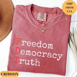 Puede incluir: Camiseta color carmes&iacute; con las palabras "Freedom Democracy Truth" impresas en blanco. La letra "D" es roja. Un collar plateado con un colgante est&aacute; sobre la camiseta. La camiseta es de Comfort Colors.