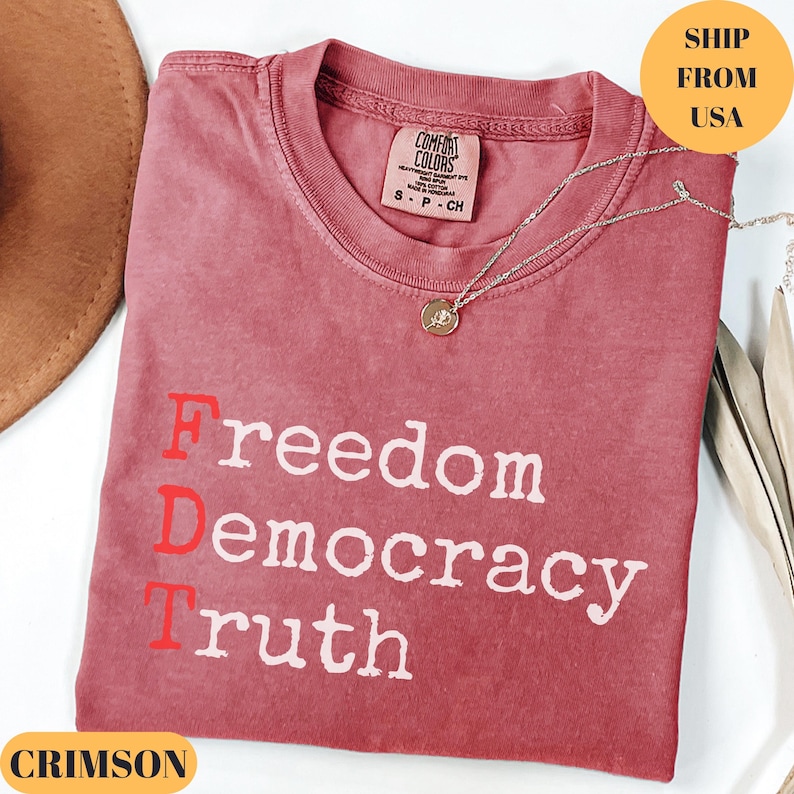 Puede incluir: Camiseta color carmes&iacute; con las palabras "Freedom Democracy Truth" impresas en blanco. La letra "D" es roja. Un collar plateado con un colgante est&aacute; sobre la camiseta. La camiseta es de Comfort Colors.