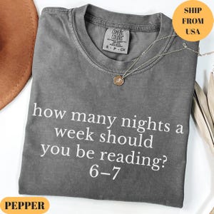 Puede incluir: Una camiseta gris Comfort Colors con el texto "how many nights a week should you be reading? 6-7" en blanco. Un collar plateado con un colgante est&aacute; sobre la camiseta. La camiseta est&aacute; doblada y tiene una etiqueta que dice "S-P-CH".