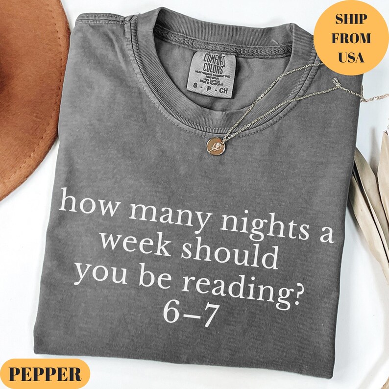 Puede incluir: Una camiseta gris Comfort Colors con el texto "how many nights a week should you be reading? 6-7" en blanco. Un collar plateado con un colgante est&aacute; sobre la camiseta. La camiseta est&aacute; doblada y tiene una etiqueta que dice "S-P-CH".