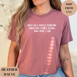 May include: Heather mauve t-shirt with the text "ONCE IN A WHILE SOMEONE AMAZING COMES ALONG, AND HERE I AM" in white. The shirt is a solid color and has a crew neck.