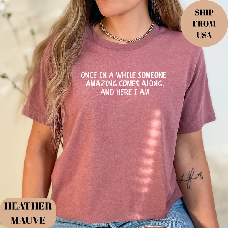 May include: Heather mauve t-shirt with the text "ONCE IN A WHILE SOMEONE AMAZING COMES ALONG, AND HERE I AM" in white. The shirt is a solid color and has a crew neck.