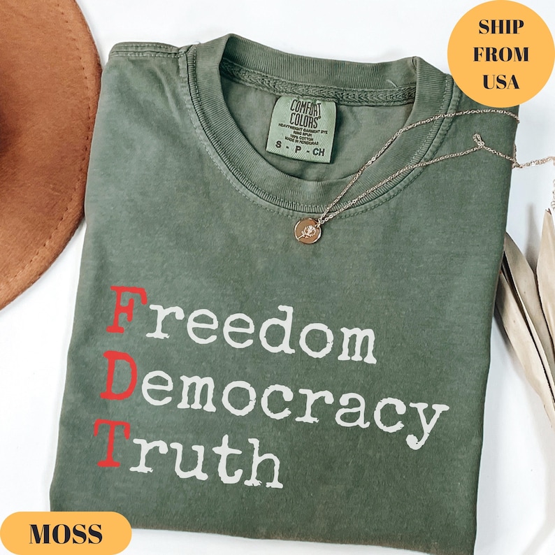 Puede incluir: Una camiseta verde musgo con las palabras "Freedom Democracy Truth" impresas en blanco y rojo. Un collar de oro con un colgante descansa sobre la camisa. La camisa est&aacute; doblada y tiene una peque&ntilde;a etiqueta.