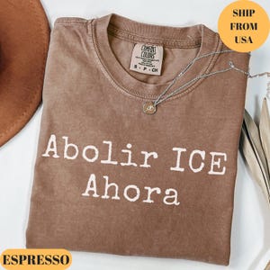 Könnte beinhalten: Espressofarbenes T-Shirt mit dem Text "Abolir ICE Ahora" in Weiß. Das Shirt hat einen Rundhalsausschnitt und ein Comfort Colors-Etikett. Eine silberne Halskette mit Anhänger liegt über dem Shirt.
