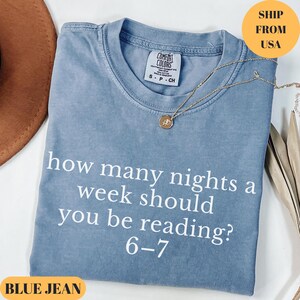 Puede incluir: Una camiseta azul vaquera con el texto "how many nights a week should you be reading? 6-7" en blanco. Un collar dorado con un colgante est&aacute; drapeado sobre la camiseta. La camiseta est&aacute; doblada sobre una superficie.