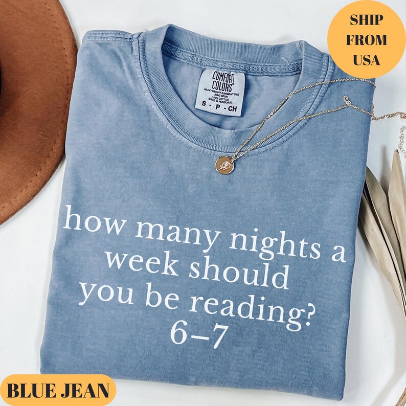 Puede incluir: Una camiseta azul vaquera con el texto "how many nights a week should you be reading? 6-7" en blanco. Un collar dorado con un colgante est&aacute; drapeado sobre la camiseta. La camiseta est&aacute; doblada sobre una superficie.