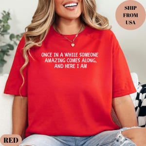 May include: A red t-shirt with the text "ONCE IN A WHILE SOMEONE AMAZING COMES ALONG, AND HERE I AM" in white. The shirt is a solid red color and has short sleeves. The shirt is a casual style.