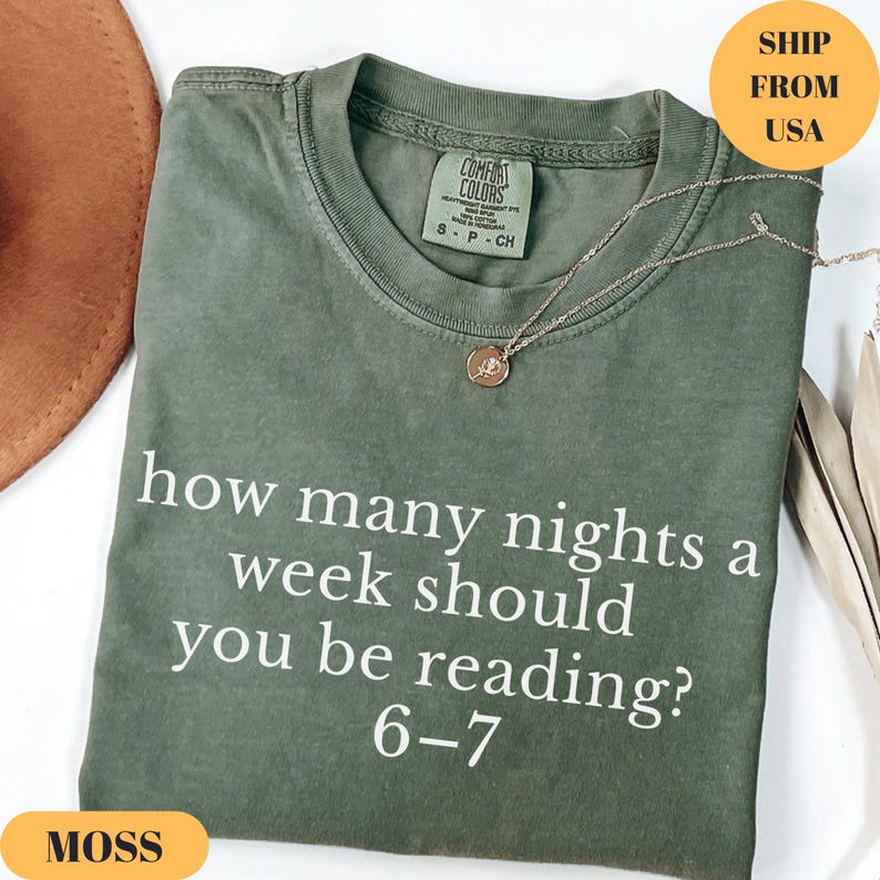 Puede incluir: Una camiseta verde musgo con el texto "how many nights a week should you be reading? 6-7" en blanco. La camiseta tiene cuello redondo y mangas cortas. Un collar dorado con un colgante est&aacute; sobre la camiseta.