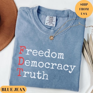 May include: A blue jean colored t-shirt with the words "Freedom Democracy Truth" printed in white and red. A gold necklace with a pendant is draped across the shirt. The shirt is folded on a white surface.
