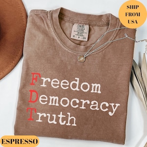 Puede incluir: Camiseta color espresso con las palabras "Freedom Democracy Truth" impresas en blanco y rojo. La camiseta tiene cuello redondo y una peque&ntilde;a etiqueta en el cuello. Un collar plateado con un colgante est&aacute; drapeado sobre la camiseta.
