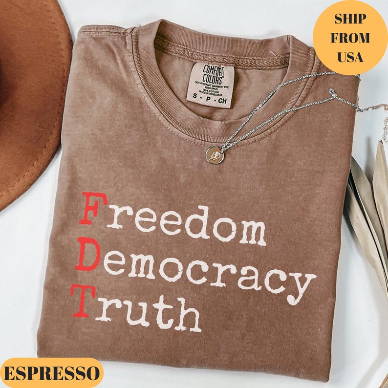 Puede incluir: Camiseta color espresso con las palabras "Freedom Democracy Truth" impresas en blanco y rojo. La camiseta tiene cuello redondo y una peque&ntilde;a etiqueta en el cuello. Un collar plateado con un colgante est&aacute; drapeado sobre la camiseta.