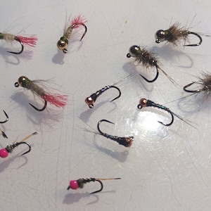 Può includere: Una collezione di mosche da pesca con vari design. Alcune mosche hanno teste color oro, corpi grigi e accenti rossi o marroni. Altre presentano perline rosa e ami neri. Le mosche sono disposte su una superficie chiara.