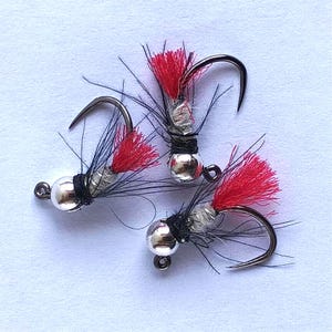 May include: Three fishing flies with silver bead heads, black thread, and red and black feather accents. The hooks are curved and made of dark metal. The flies are arranged on a white surface.