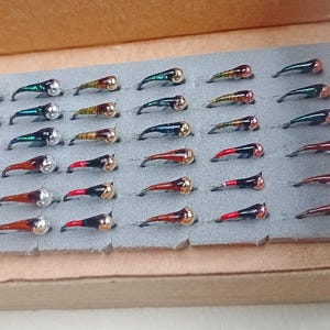 Puede incluir: Una colección de treinta y cinco moscas de pesca dispuestas en siete filas sobre un inserto de espuma gris dentro de una caja de cartón. Cada mosca presenta un anzuelo curvo, una cabeza de cuenta metálica y un cuerpo de colores variados.