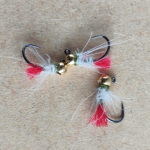 May include: Three fishing flies with gold-colored heads, black hooks, and white, red, and green accents. The flies are designed for catching fish. The flies are arranged on a light brown surface.