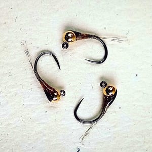 May include: Three fishing flies featuring curved black metal hooks, shiny gold-coloured bead heads, and dark brown tapered bodies with thin, light-coloured synthetic fibres extending from the rear of each hook to mimic insect tails.